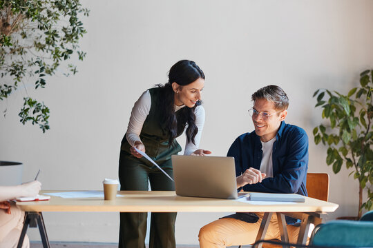 Business professionals collaborating at office desk, discussing project on laptop and documents, sharing ideas during meeting, working together in modern workspace