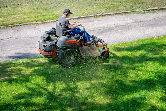 Man riding professional zero turn lawn mower machine cutting green grass in park. Gardener operating riding lawnmower for lawn care maintenance. No faces