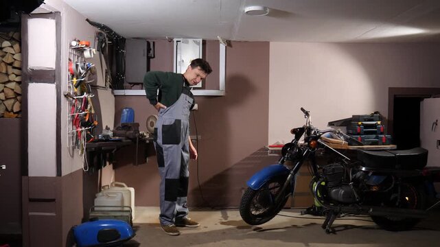Male mechanic in overalls feeling acute lower back pain while standing next to a vintage motorcycle in a home workshop, highlighting health issues from physical labor