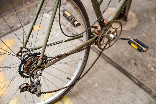 Detailed close up of a bicycle chain, rear gear cassette, and pedals parked on a city sidewalk. Urban transportation concept showing metal components, wheel spokes, and mechanical drivetrain system.