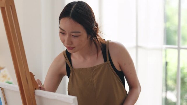 Creative young Asian woman artist painting on canvas with brush in bright home studio. Relaxed female hobbyist enjoying peaceful indoor leisure time, expressing imagination for art.