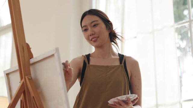 Creative young Asian woman artist painting on canvas with brush in bright home studio. Relaxed female hobbyist enjoying peaceful indoor leisure time, expressing imagination for art.