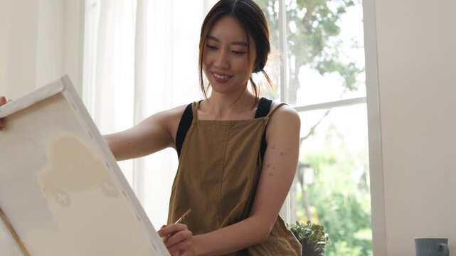 Creative young Asian woman artist painting on canvas with brush in bright home studio. Relaxed female hobbyist enjoying peaceful indoor leisure time, expressing imagination for art.