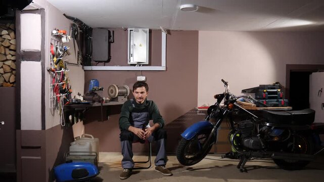 Young man sitting on a chair, pausing from his work on a vintage motorcycle in a cluttered private garage, enjoying a cold drink, surrounded by various tools and equipment