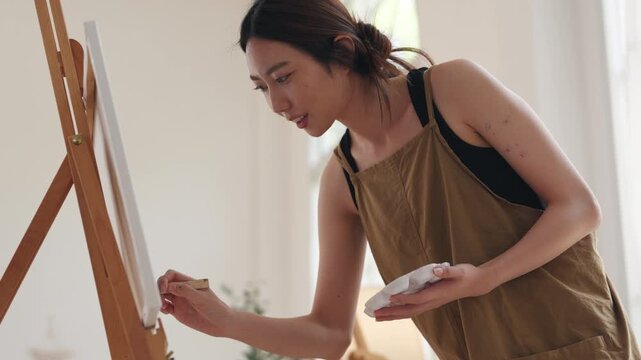 Creative young Asian woman artist painting on canvas with brush in bright home studio. Relaxed female hobbyist enjoying peaceful indoor leisure time, expressing imagination for art.