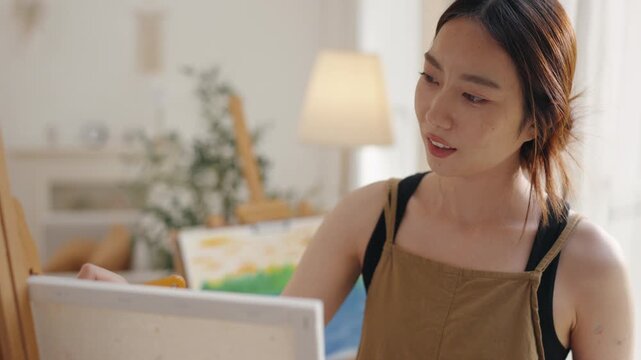 Creative young Asian woman artist painting on canvas with brush in bright home studio. Relaxed female hobbyist enjoying peaceful indoor leisure time, expressing imagination for art.