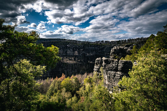 View of Pravcicka Brana sandstone arch formation surrounded by lush pine forests and dramatic rocky cliffs under a cloudy blue sky in Hrensko, Usti nad Labem Region, Czechia.