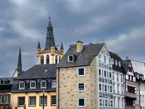 View of the historic St. Gangolf church tower rising above traditional timber-framed buildings and stone facades under a cloudy sky in Trier, Rheinland-Pfalz, Germany.