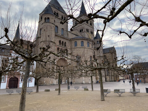 View of Trier Cathedral with its historic stone facade, arched doorways, and bare trees in the foreground under a cloudy sky in Trier, Rheinland-Pfalz, Germany.