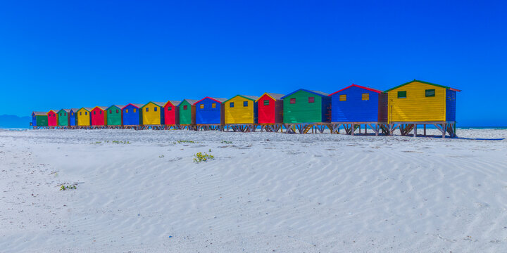 View of a row of colorful wooden beach huts standing on the white sand of Muizenberg beach under a clear blue sky in South Africa.