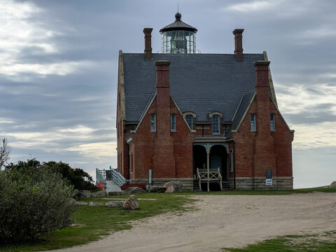View of the historic Block Island Southeast Light featuring a red brick facade and slate roof under a cloudy sky in New Shoreham, Rhode Island, United States.