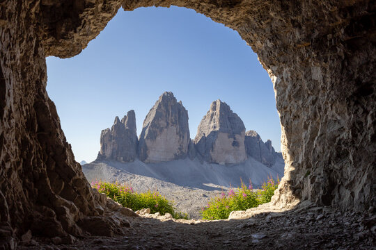 View of Tre Cime di Lavaredo framed by a natural rock cave opening with rugged stone textures, vibrant wildflowers, and clear blue sky in Auronzo di Cadore, Veneto, Italy.