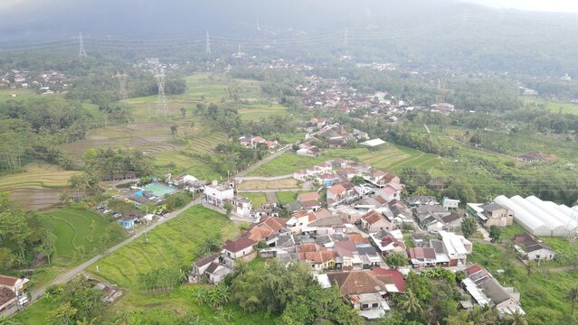 Ungaran, Central Java, Indonesia - 09 April 2021: Aerial view of a rural village nestled among lush green rice terraces and rolling hills under a soft overcast sky.