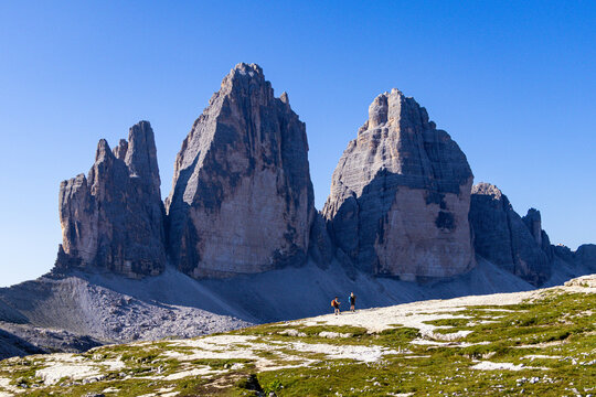 View of Tre Cime di Lavaredo with hikers walking on a rocky trail under a clear blue sky in Auronzo di Cadore, Veneto, Italy.