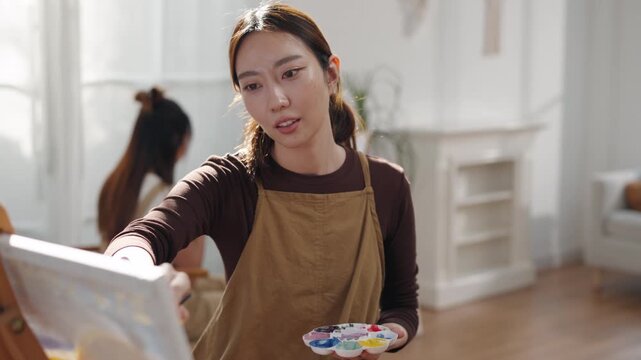 Creative young Asian woman artist painting on canvas with brush in bright home studio. Relaxed female hobbyist enjoying peaceful indoor leisure time, expressing imagination for art.