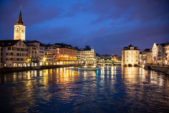 View of the Limmat River at dusk with the historic St. Peter Church tower, illuminated buildings, and reflections on the water in Zurich, Zurich, Switzerland.