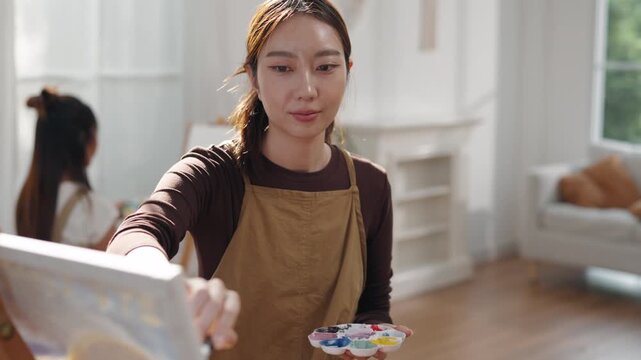 Creative young Asian woman artist painting on canvas with brush in bright home studio. Relaxed female hobbyist enjoying peaceful indoor leisure time, expressing imagination for art.