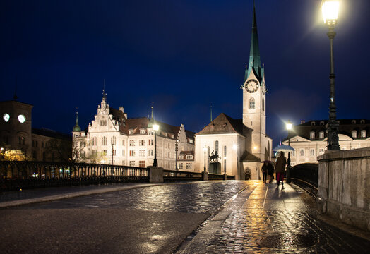 View of Fraumunster church illuminated at night with street lamps, cobblestone bridge, and pedestrians in Zurich, Zurich, Switzerland.