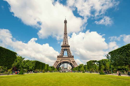View of the Eiffel Tower rising above manicured green lawns and trimmed trees under a bright blue sky with fluffy white clouds in Paris, Ile-de-France, France.