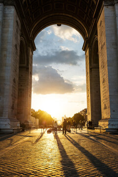 View of the Arc de Triomphe at sunset with people walking on the cobblestone pavement and long shadows stretching across the ground in Paris, Ile-de-France, France.