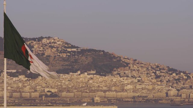 Aerial view of the Algerian flag waving in the wind with the coastal city of Algiers and hillside buildings in the background in Algeria.