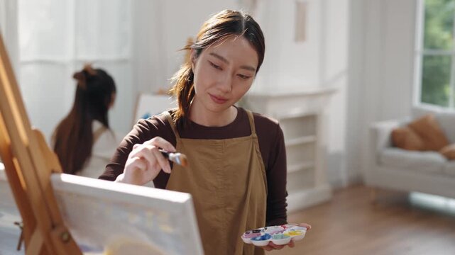 Creative young Asian woman artist painting on canvas with brush in bright home studio. Relaxed female hobbyist enjoying peaceful indoor leisure time, expressing imagination for art.