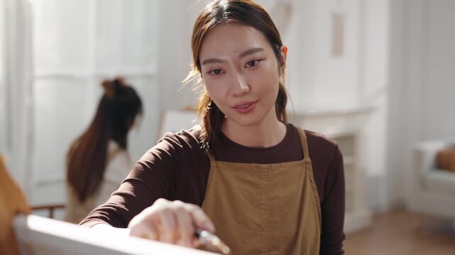 Creative young Asian woman artist painting on canvas with brush in bright home studio. Relaxed female hobbyist enjoying peaceful indoor leisure time, expressing imagination for art.