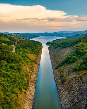 Aerial view of the Presa de Taveras reservoir surrounded by lush green hills and steep rocky embankments under a soft sunset sky in Presa de Taveras, La Vega, Dominican Republic.