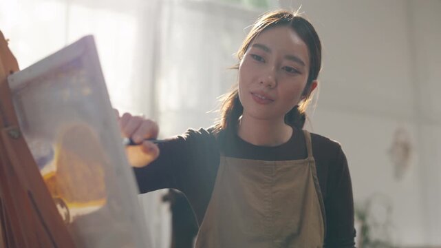 Creative young Asian woman artist painting on canvas with brush in bright home studio. Relaxed female hobbyist enjoying peaceful indoor leisure time, expressing imagination for art.