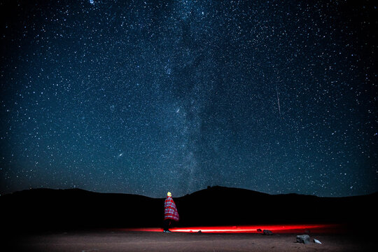 View of a person wrapped in a plaid blanket standing in the desert under a vast starry night sky with the Milky Way visible and a red light glowing on the ground in Algeria.