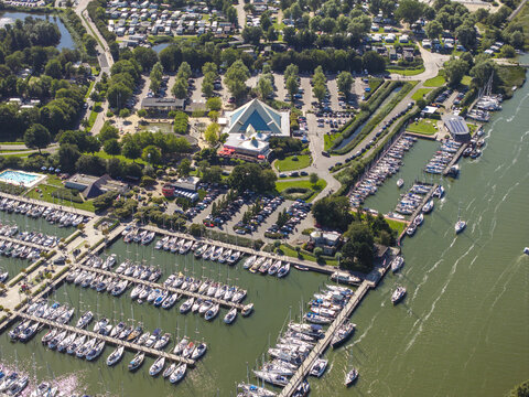 Aerial view of De Holle Poarte marina with numerous sailboats docked in the harbor, adjacent parking lots, and surrounding green trees in Makkum, Friesland, Netherlands.