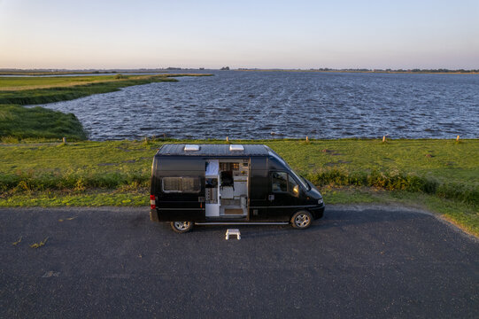 Aerial view of a black camper van parked on a paved area beside a lake with a grassy bank and calm water in Hemelum, Friesland, Netherlands.