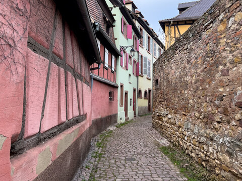 View of a narrow cobblestone street lined with traditional half-timbered houses and a historic stone wall in the old town of Colmar, Grand Est, France.