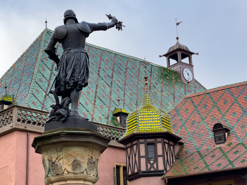 View of the bronze statue of Admiral Bruat standing before the historic Koifhus with its iconic colorful tiled roof and clock tower in Colmar, Grand Est, France.