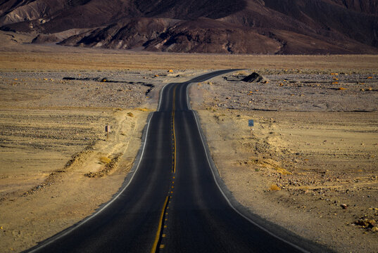 View of a long, winding asphalt road stretching through the arid desert landscape of Death Valley National Park, with rugged mountains in the background, Death Valley, California, United States.
