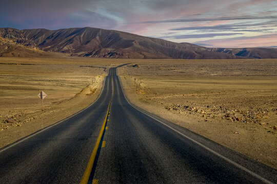 View of a long asphalt road stretching through the arid desert landscape of Death Valley National Park's backcountry under a soft sunset sky in Death Valley, California, United States.