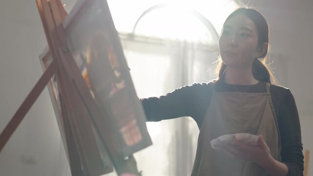 Creative young Asian woman artist painting on canvas with brush in bright home studio. Relaxed female hobbyist enjoying peaceful indoor leisure time, expressing imagination for art.