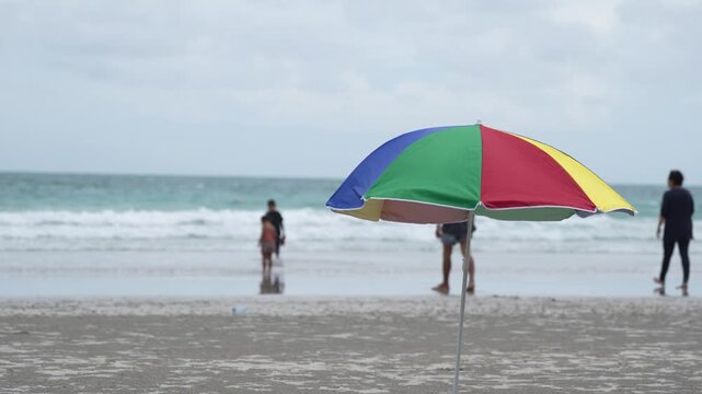 Beach umbrella and parasol on seaside shoreline, ocean waves over wet sand, summer vacation travel scene with family silhouette and child playing in gentle surf under cloudy sky, calm mood, copy space
