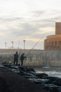 Casablanca, Casablanca-Settat, Morocco - 15 November 2025: View of two fishermen standing on the rocky breakwater near the Hassan II Mosque with the Atlantic Ocean in the background.