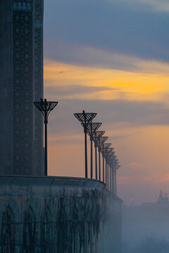 Casablanca, Morocco - 15 November 2025: View of the Hassan II Mosque at sunset, featuring ornate stone walls and a row of decorative street lamps silhouetted against a golden sky.