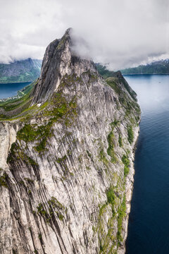 Aerial view of the dramatic Segla mountain peak with steep cliffs and lush green vegetation rising above the deep blue waters of the fjord in Senja, Troms, Norway.