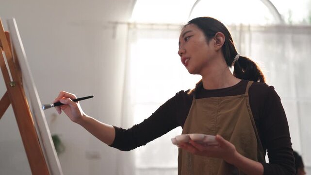 Creative young Asian woman artist painting on canvas with brush in bright home studio. Relaxed female hobbyist enjoying peaceful indoor leisure time, expressing imagination for art.