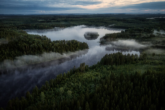 Aerial view of a serene lake surrounded by dense coniferous forests with morning mist hovering over the water in Kangasniemi, South Savo, Finland.