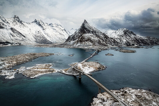 Aerial view of the Fredvang Bridges connecting islands in the Lofoten archipelago, surrounded by snow-capped mountains, icy blue waters, and dramatic winter clouds in Lofoten, Nordland, Norway.