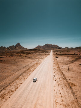 Namibia Desert, Namibia - 10 September 2023: View of a white car driving along a long, straight dirt road through the vast, arid landscape of the Namib Desert with distant mountains.