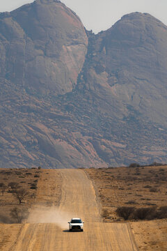 Namib Desert, Namibia - 10 September 2023: View of a white car driving on a dusty gravel road with the granite peaks of Spitzkoppe rising in the background.