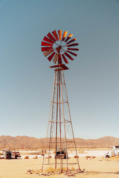 Namib Desert, Namibia - 10 September 2023: View of a rustic metal windmill in the arid desert landscape with vintage abandoned vehicles and distant mountains under a clear sky.