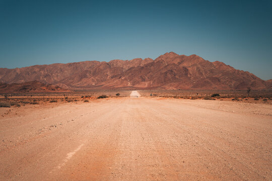 Namibia Desert, Namibia - 10 September 2023: View of a long, dusty gravel road stretching towards the rugged, sun-drenched mountains of the Namib Desert under a clear blue sky.