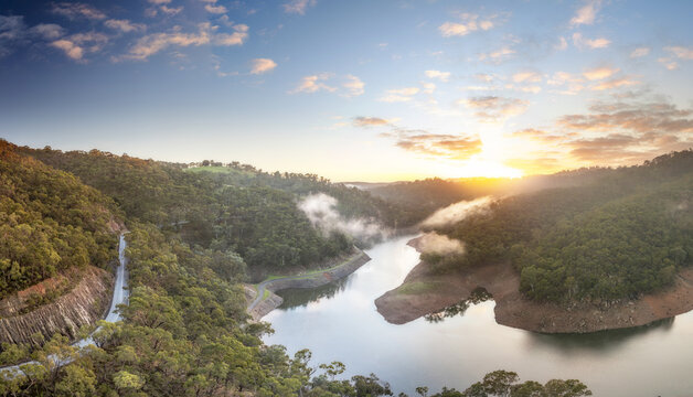 Aerial view of Kangaroo Creek Reservoir at sunrise with mist over the water, surrounded by eucalyptus forests and a winding road in Adelaide Hills Council, South Australia, Australia.