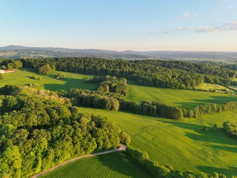Aerial view of the Rhon Mountains landscape featuring lush green meadows, dense forests, and rolling hills under soft golden hour light in Fulda, Hessen, Germany.